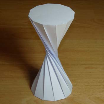 Folded paper model of an hourglass-shaped hyperboloid on a tabletop.